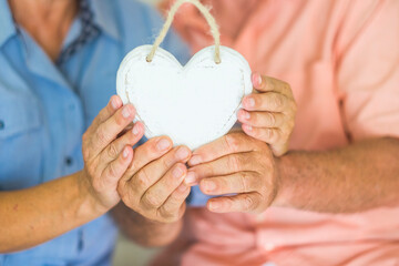 Close up of couple old hands holding white wooden heart to represent love and relationship. Heart...