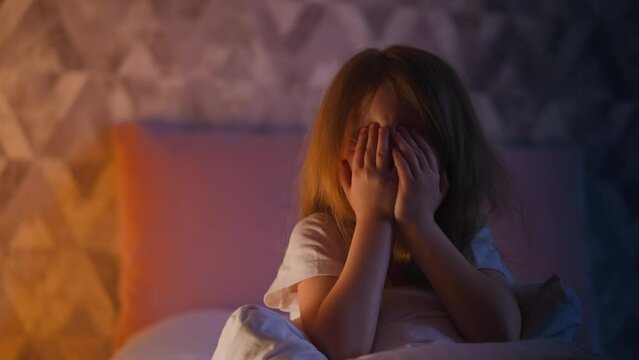 Emotional little girl with blonde hair flings arms up to hide face sitting on bed in shaded bedroom with gentle orange lamp light and shadows slow motion