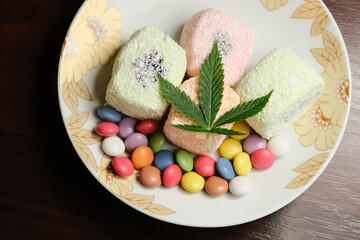 Marijuana sweet dessert. Turkish delight and colorful candy confetti with green cannabis leaf close up. Layout of weed food with CBD oil on white plate. Top view, flat lay, wooden background.