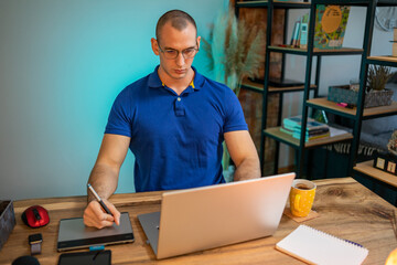 Young handsome man working from home using a laptop and a graphics tablet for his freelance project