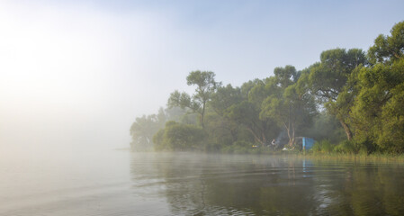 Fototapeta premium Mystical landscape. Fog in the early morning on the river. The trees near the water are illuminated by the rays of the rising sun.