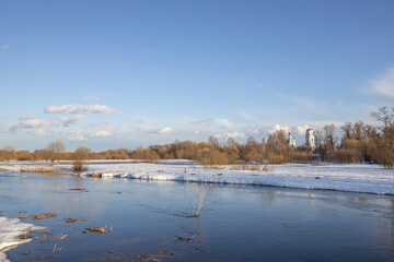 March sunny day by the river. A picturesque landscape, early spring, a river with snow-covered banks, dry grass and bushes. Church in the background. The first thaws, the snow is melting