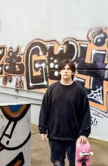 Teenager with skateboard standing in front of a graffitied wall