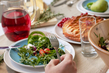 Close-up view of unrecognizable person sitting at dining table and having appetizing lunch: fresh arugula salad, homemade pie and berry kompot