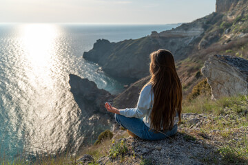 Woman tourist enjoying the sunset over the sea mountain landscape. Sits outdoors on a rock above the sea. She is wearing jeans and a blue hoodie. Healthy lifestyle, harmony and meditation