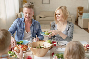 Happy Caucasian family with children sitting at dining table at home, eating tasty dinner and enjoying time together, mother offering fresh fruit