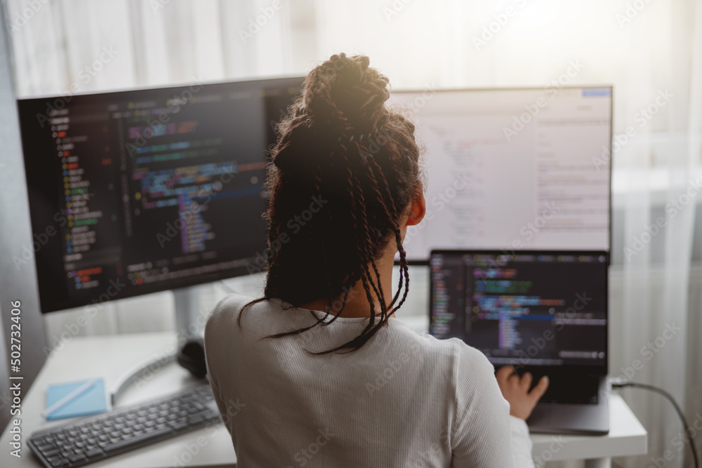 Wall mural rear of female web expert typing on computer looking at monitors while sitting indoors