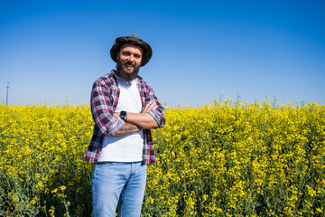 Naklejka na ściany i meble Portrait of happy and successful farmer who is standing by his rapeseed field. Rapeseed plantation in bloom.