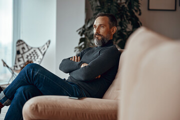 Side view of serious man with arms crossed sitting in cosy armchair