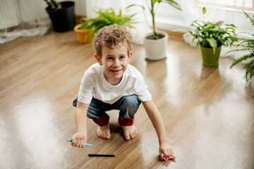 The boy is squatting on the floor in the room, holding a marker in his hand, wiping the floor with a napkin