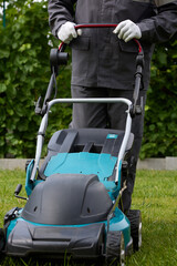 Vertical photo of a man in overalls and white gloves mowing green grass in the garden with a lawn mower. A worker in coveralls uses a modern lawnmower in the garden. Professional lawn care service.