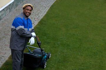 Top view of an African man in overalls mowing green grass in a modern garden with a lawn mower. A...