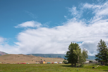 Dramatic alpine view from trees to small houses in sunlit green meadow against high mountain range under clouds in blue sky. Scenic mountain landscape at changeable weather. Village in high mountains.