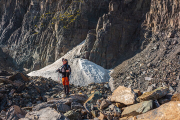 Hiker with trekking poles stands on moraine against rocks with glacier in bright sunlight. Man in sunglasses in bright sun near sunlit rocky mountain wall. Tourist in high mountains in sunny day.