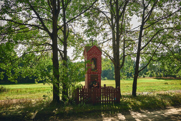 A red brick wayside shrine standing by a dirt road in the shade of trees. Tuchola Forest, Poland - obrazy, fototapety, plakaty