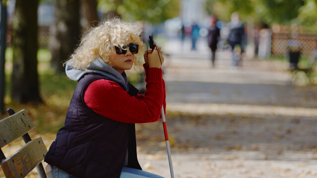 Young caucasian blonde woman with vision disability sitting on a bench in a park, keeping her hands on white cane. Life with disability concept. High quality photo
