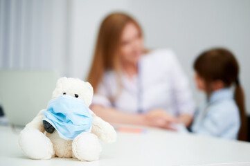 Selective focus. Teddy bear with medical mask in hospital with female caucasian doctor and child
