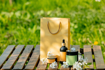 Cosmetic bottle and jar next to shopping bag on wooden table in blooming tree garden