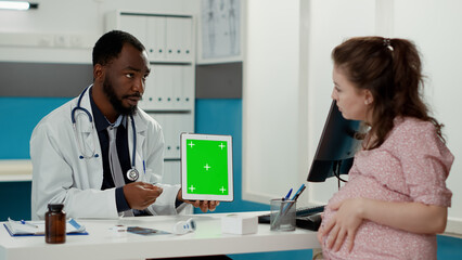 Male doctor showing tablet with greenscreen on display to pregnant patient at office desk. Medic using isolated mockup template with chroma key background and blank copy space. Tripod shot.