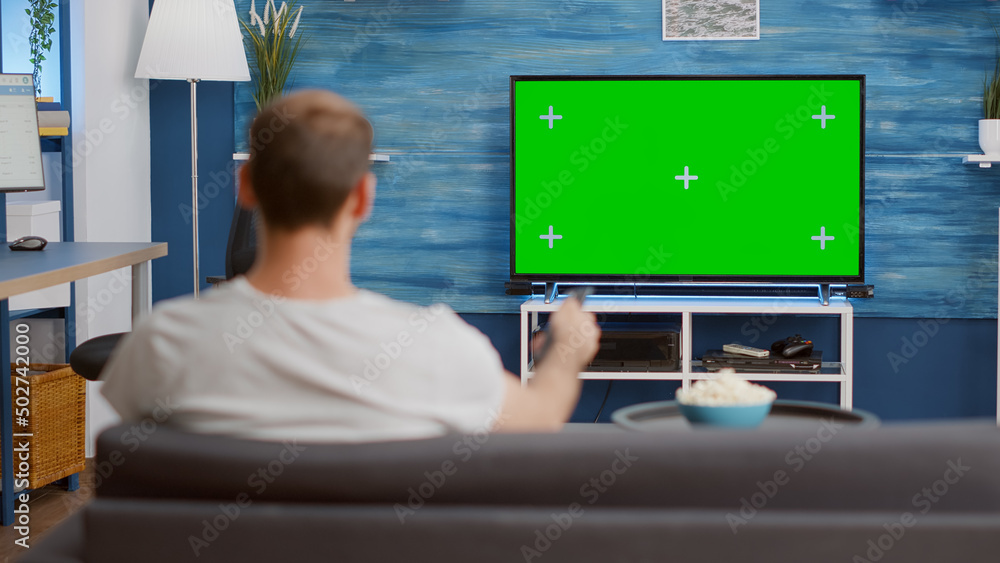 Poster man looking at green screen on tv and switching channels sitting on couch in modern living room with