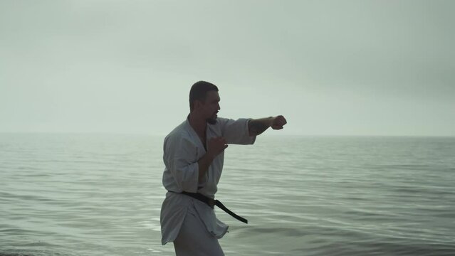 Strong man practicing taekwondo near ocean against cloudy sky in slow motion.