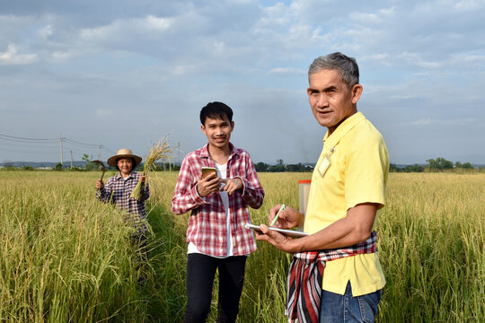 Portrait of Asian senior surveyer who wears yellow shirt and holds pen and notebook infront of asian young boy and asian elderly woman to survey and store rice growing informaton .