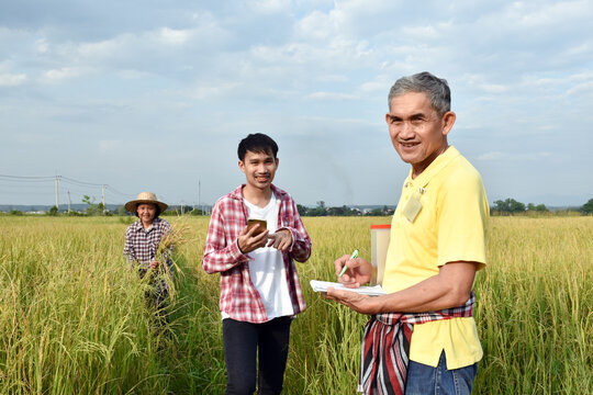 Portrait of Asian senior surveyer who wears yellow shirt and holds pen and notebook infront of asian young boy and asian elderly woman to survey and store rice growing informaton .