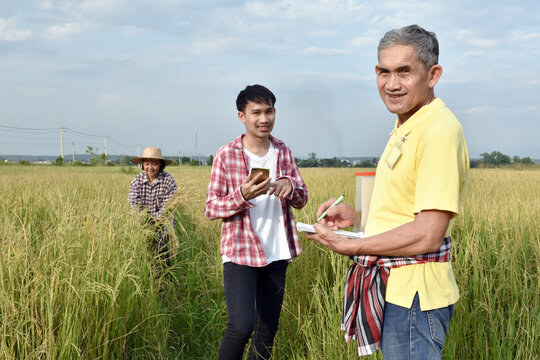 Portrait of Asian senior surveyer who wears yellow shirt and holds pen and notebook infront of asian young boy and asian elderly woman to survey and store rice growing informaton .