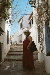 Fototapeta premium young pregnant woman with red dress and hat walking in traditional spanish town with white buildings and clear blue sky on a sunny day, with traditional spanish architecture