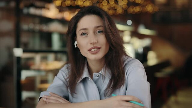 Portrait of confident young woman in wireless earphones sitting at cafe and talking on camera during video call. Concept of people, interview and vlogging.
