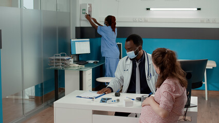 Naklejka premium African american medic and expectant woman with face mask at checkup visit to give prescription medicine in bottle of pills. Doctor giving treatment to pregnant patient with baby bump.