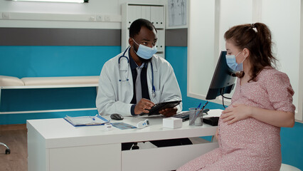 Fototapeta premium Male obstetrician using digital tablet at checkup appointment with pregnant patient in office. Doctor taking notes and doing health care consultation with woman during covid 19 pandemic.