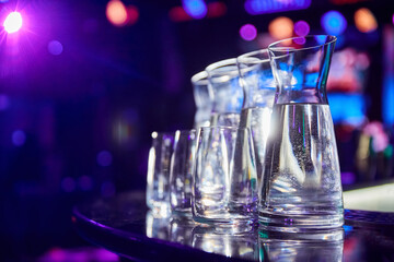 Carafes with water and glasses on the bar of a nightclub against the backdrop of multi-colored spotlights