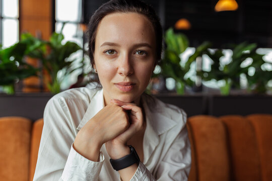 Portrait of young businesswoman or manager in white shirt leaning on hands and looking at camera in modern restaurant  