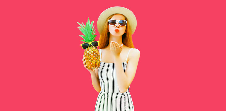 Summer portrait of beautiful young woman with pineapple blowing her red lips sending sweet air kiss wearing straw hat, striped jumpsuit on pink background