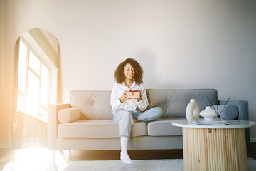 Excited african American young woman sit on couch at home unpack gift box, overjoyed girl feel...