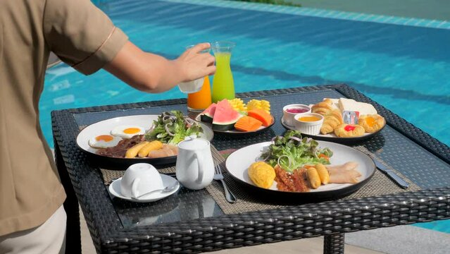 Waiter hand serving breakfast in restaurant at luxury hotel. Asian woman waitress preparing morning food and drinks on table in outdoors pool cafe in tropical resort. Travel, holiday, vacation concept
