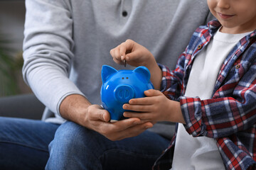 Little boy with his father putting coin into piggy bank indoors, closeup