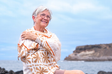 Portrait of smiling senior woman wearing eyeglasses and scarf sitting outdoors at sea enjoying good time, freedom and relax. Cloudy sky, copy space. Travel vacation retirement concept