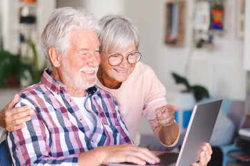 Joyful beautiful senior couple smiling at home browsing together on laptop. Active elderly Caucasian couple enjoying technology and social