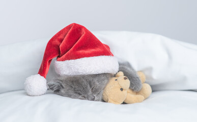 Funny kitten wearing red santa's hat sleeps under white blanket and hugs favorite toy bear