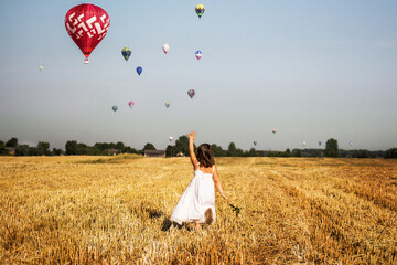 Dziewczynka biegnie po polu i macha do balonów na gorące powietrze, lot balonem nad polami w środku lata - obrazy, fototapety, plakaty