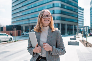 European 30s businesswoman in stylish clothes with a laptop walking outdoors near the business center