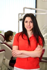 Vertical Portrait of happy beautiful female doctor, attractive pretty middle aged Caucasian Armenian ethnic dentist in red polo standing in dental office at clinic, smiling, looking at camera. 