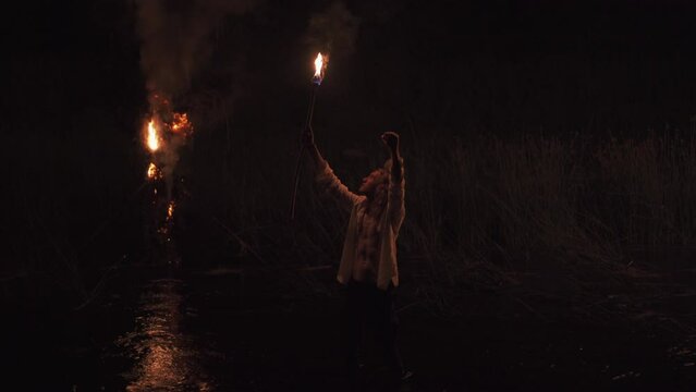 Cinematic night ritual shot with fire. Man with torch in river raises his hands