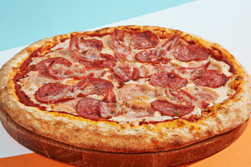 Italian pizza with salami, ham, bacon, pepperoni on coloured background. Meat pizza with sausage and bacon in minimal style on blue, orange color. American pizza delivery concept with color backdrop.