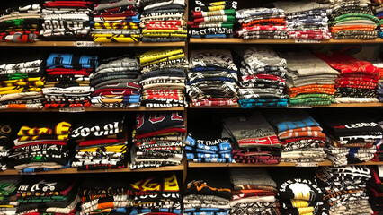 A large assortment of bright cotton T-shirts neatly folded on the shelf. Fresh T-shirts of different sizes stacked on a display case with shelves.