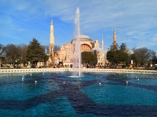 Naklejka premium Istanbul Turkey, AyaSofya Haqia Sophia Constanipole view blue water fountain in front of mosque birds in the sky, blue colored sky and water along with green treens and minaretes tourists visiting.