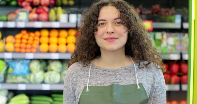 Portrait of young saleswoman in green apron standing in supermarket with shelves of fruits and vegetables on background, looking at camera and smiling.