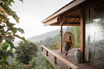Tourist woman swing on wicker rattan hang chair in the jungle, nature mountains view, hold in hands cup of tea/coffee - obrazy, fototapety, plakaty